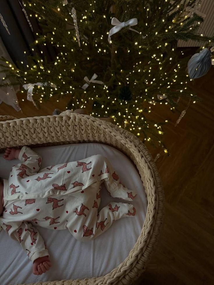 Baby in a Millie and Me romper lying in a wicker Moses basket beside a lit Christmas tree