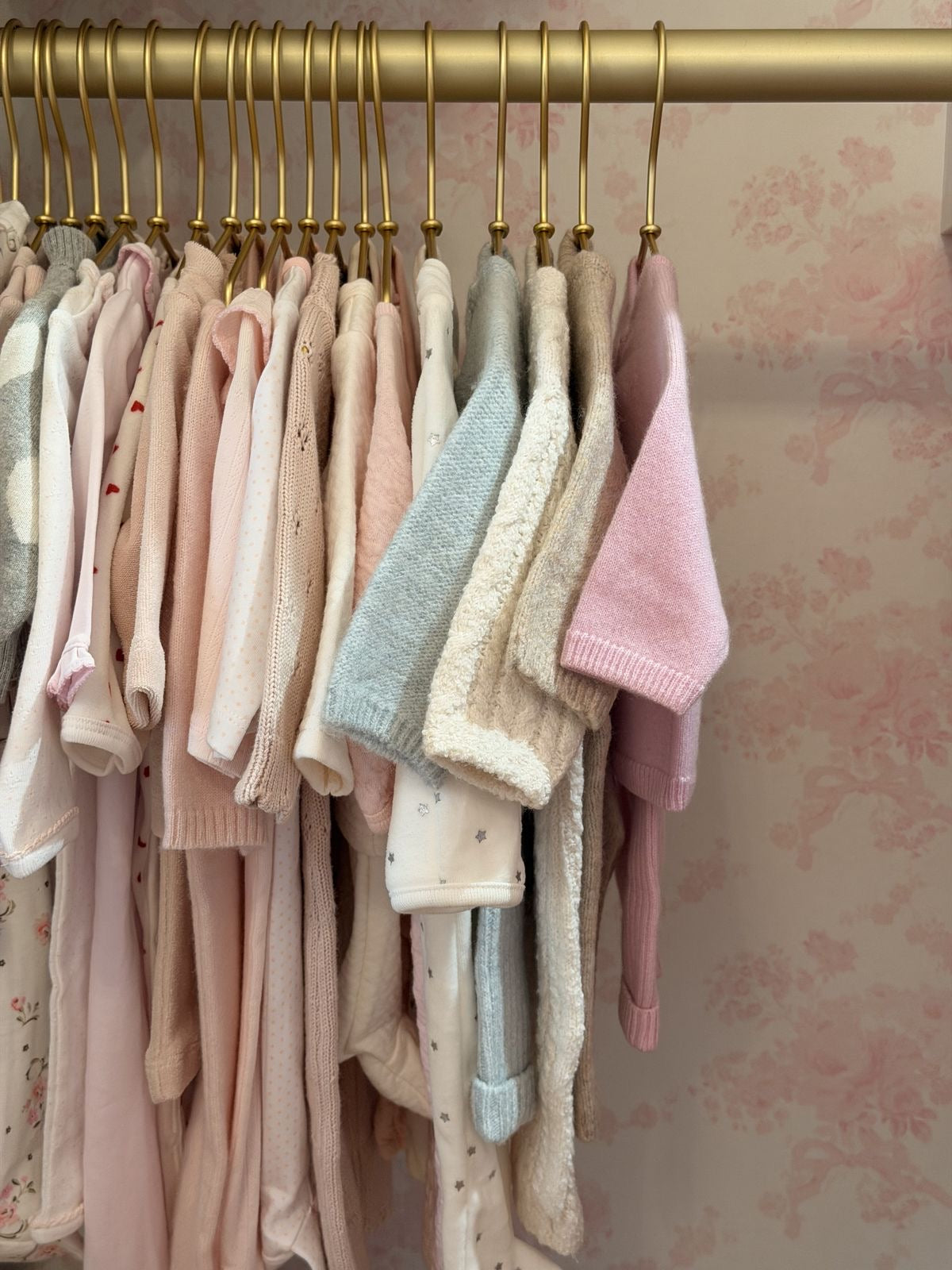 Pastel pink, cream and blue baby clothes hanging on gold hangers in a nursery, showcasing soft knitwear and newborn essentials.