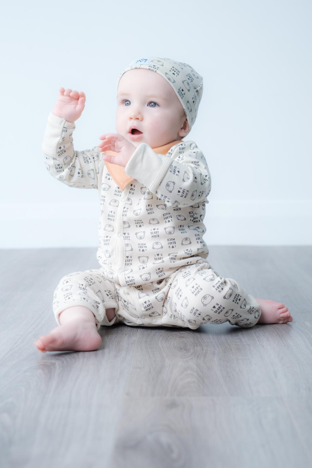 A baby sitting on a grey wood floor wearing a cream-coloured one-piece jumpsuit and matching bonnet with a small black geometric pattern, set against a plain light grey background.
