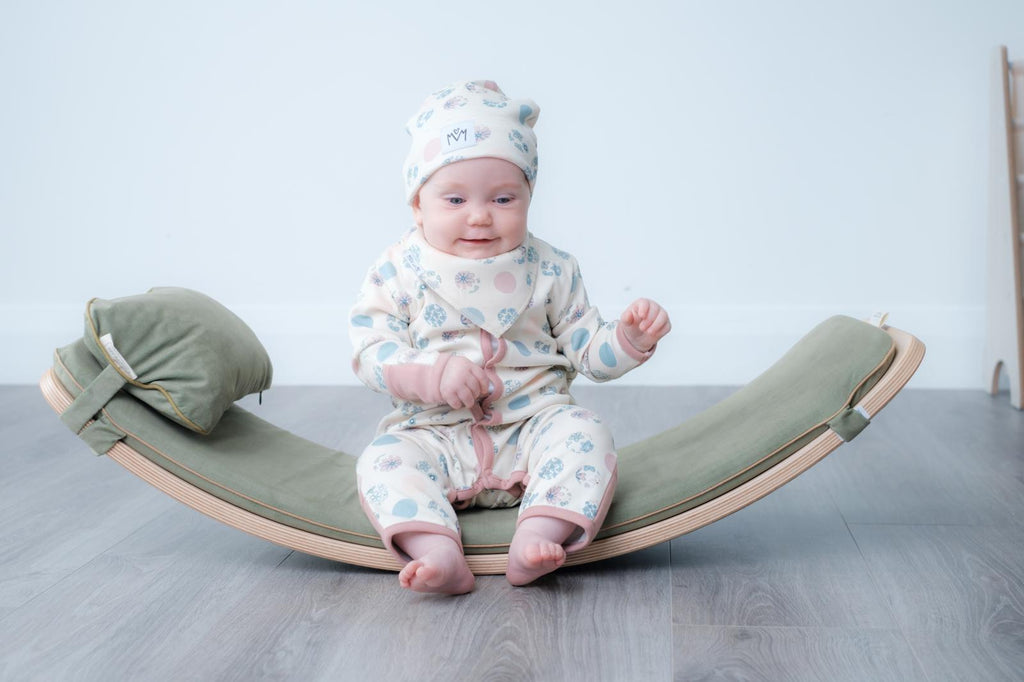 A baby wearing a light patterned footed onesie and matching hat sits centreed on a curved olive-green rocking cushion with a wooden base on a grey floor against a pale blue wall.