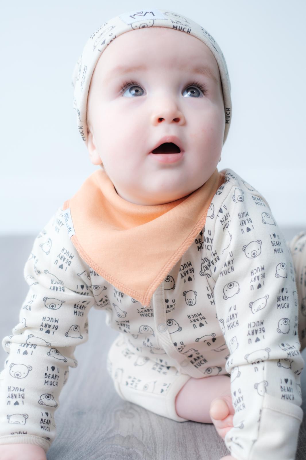 A baby wearing a cream-colored long-sleeve onesie with a black cartoon animal print, a matching beanie, and a peach-colored bib, lying on a light gray surface.
