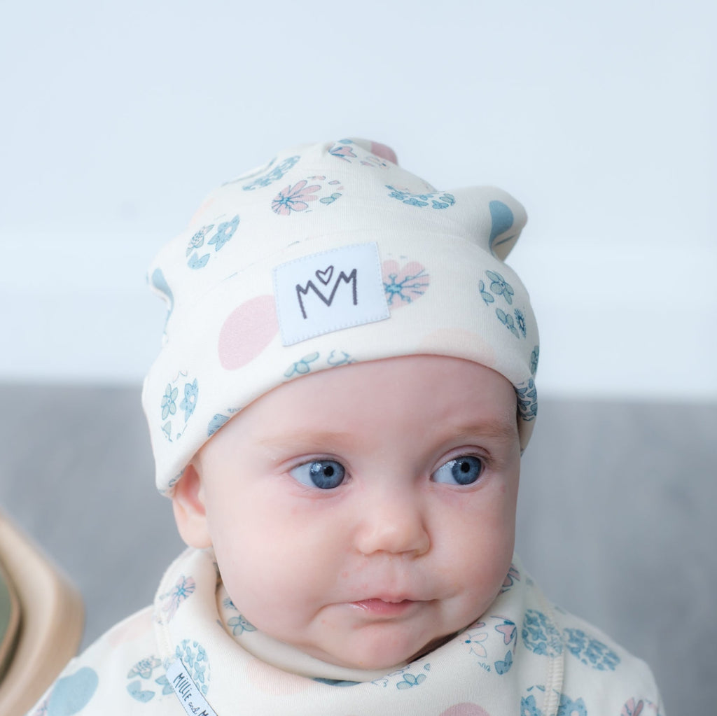 A baby wearing a cream-coloured knit beanie hat with a blue and pink floral pattern, a matching bib, and a small white patch that says 'MM' in black letters. The photo is taken in a softly lit indoor setting with a blurred background.