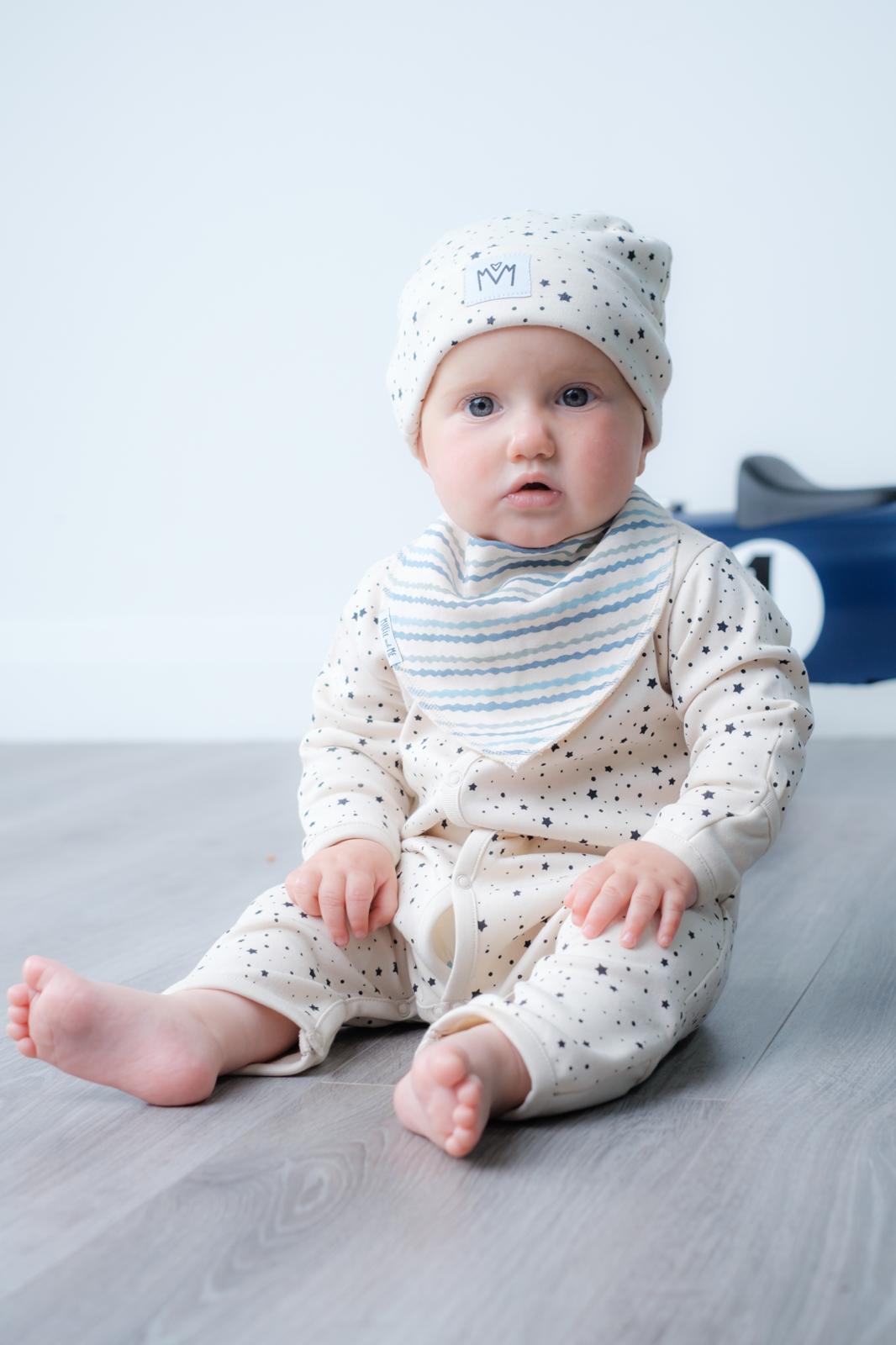 A baby sitting on a grey wooden floor wearing a cream-colored long-sleeve romper with black speckles, a striped bib, and a matching speckled bonnet.