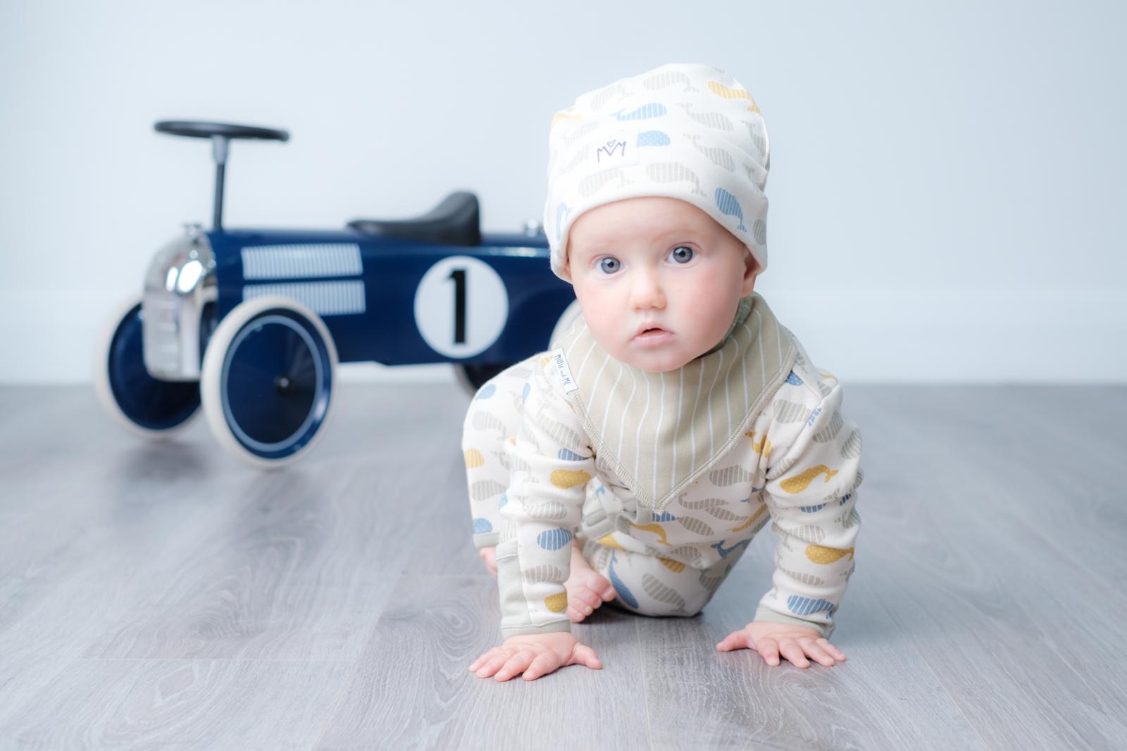 A baby crawling on a grey wooden floor wearing a cream-coloured striped onesie with blue and yellow cloud patterns and a matching beanie hat. A blue toy race car is in the background against a light grey wall.