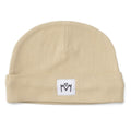 A light beige knit beanie hat with a folded cuff, featuring a small white square patch on the front with a black embroidered logo.