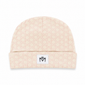 A light beige knit beanie with a repeating hexagon pattern in a soft pink, featuring a white rectangular patch on the folded cuff with a black stylized logo.