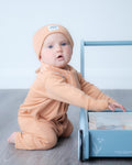 A baby wearing a peach-colored one-piece romper and matching beanie hat sits on a gray wood floor next to a light blue wooden toy cart.
