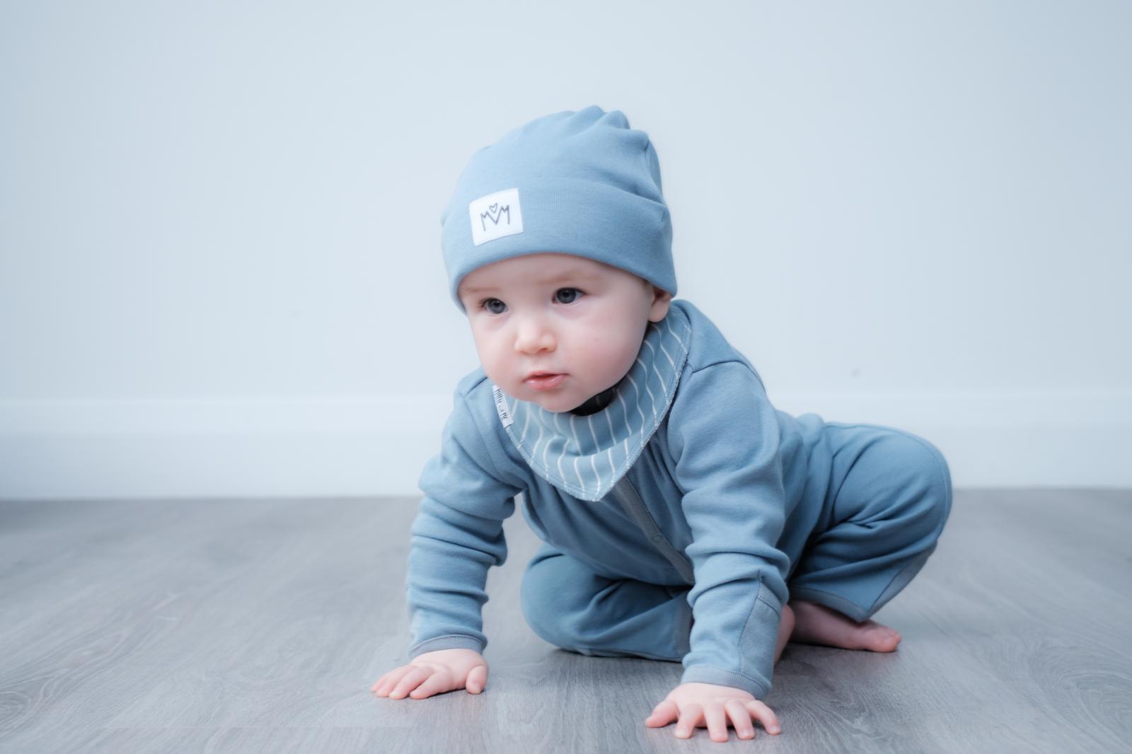 A baby crawling on a grey wood floor against a light grey wall, wearing a light blue long-sleeve one-piece outfit, a matching beanie hat, and a striped bib.