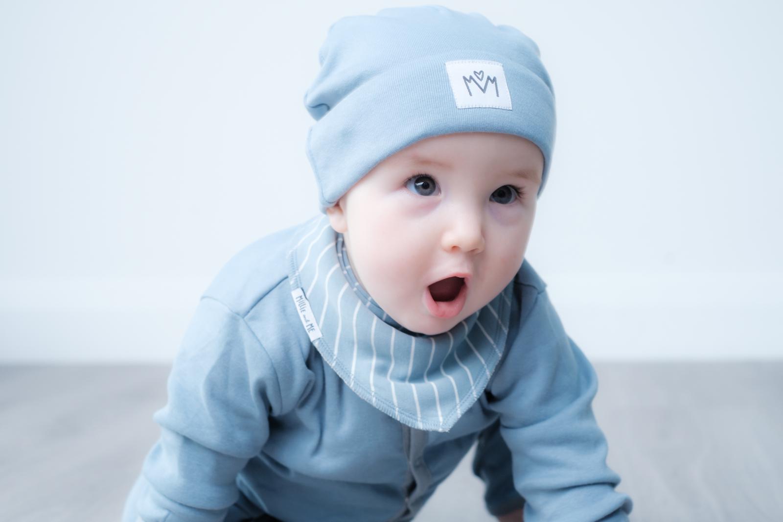 A baby crawling on a light grey floor wearing a light blue beanie with a small white square patch, a matching light blue top, and a white striped bib.