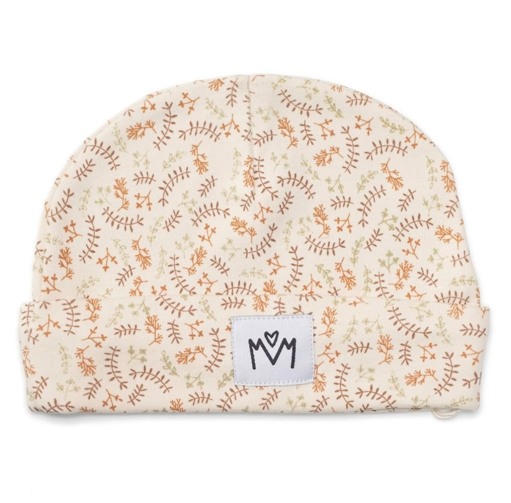A baby beanie hat featuring a delicate repeating pattern of brown and green botanical line drawings on a warm cream background. A white rectangular brand patch with a black stylised logo is sewn onto the front cuff.