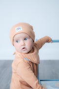 A baby wearing a peach-coloured knit beanie with a small white patch and a matching striped bib, posed next to a blue stroller against a light grey background.