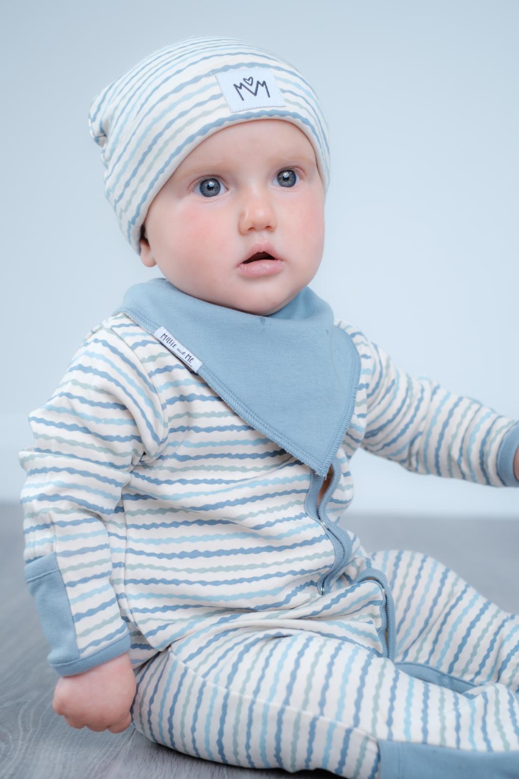 A baby wearing a light blue and white striped footed sleeper with a matching beanie and a solid light blue bib, sitting against a pale blue-grey background.