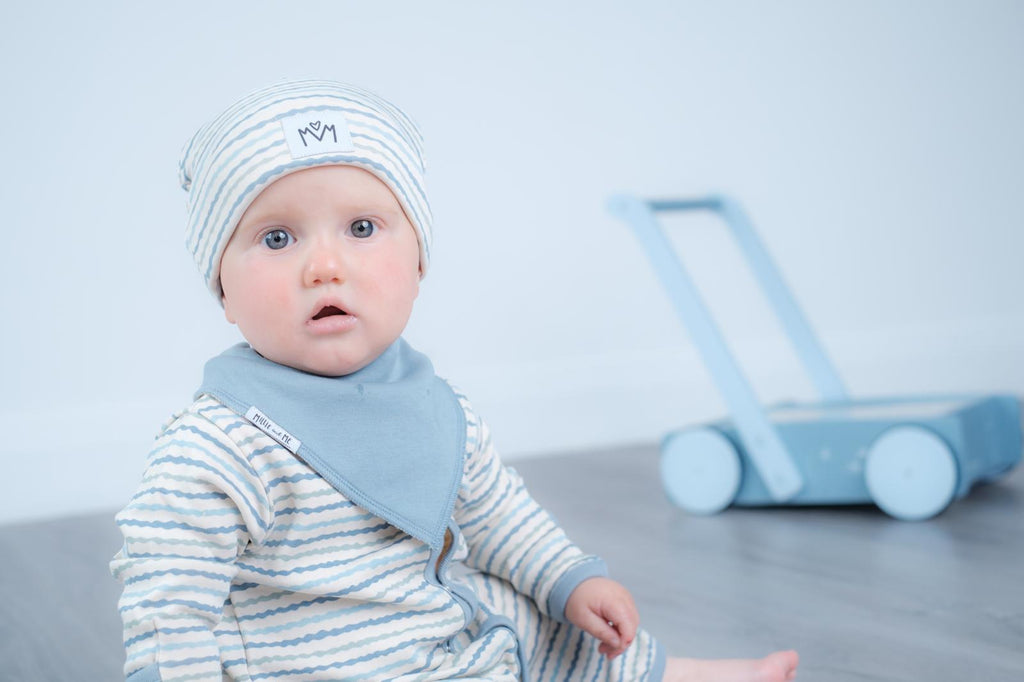 A baby wearing a light blue and white striped long-sleeve bodysuit with a matching bib and beanie hat, sitting in a studio setting with a toy walker in the background.