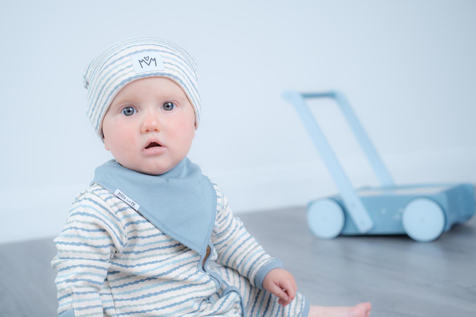 A baby wearing a light blue and white striped long-sleeve bodysuit with a matching bib and beanie hat, sitting in a studio setting with a toy walker in the background.