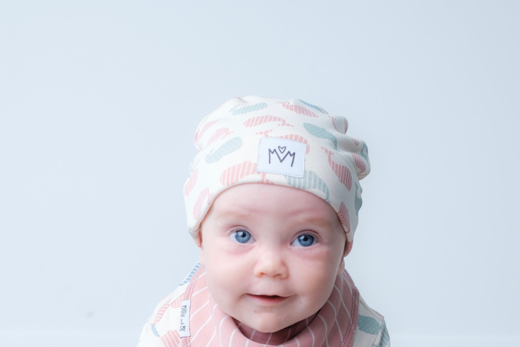 A baby wearing a soft knit beanie hat with a whale pattern in pink, blue, and grey on a cream background, along with a matching striped bib. The hat features a white patch with the brand logo.