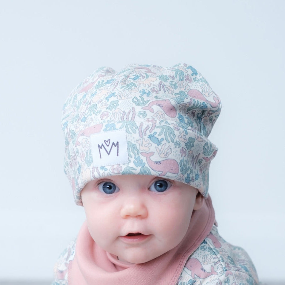 A close-up of a baby wearing a light blue and pink patterned knit beanie with a white logo patch, paired with a soft pink neck warmer, against a clean light gray background.