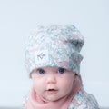 A close-up of a baby wearing a light blue and pink patterned knit beanie with a white logo patch, paired with a soft pink neck warmer, against a clean light gray background.