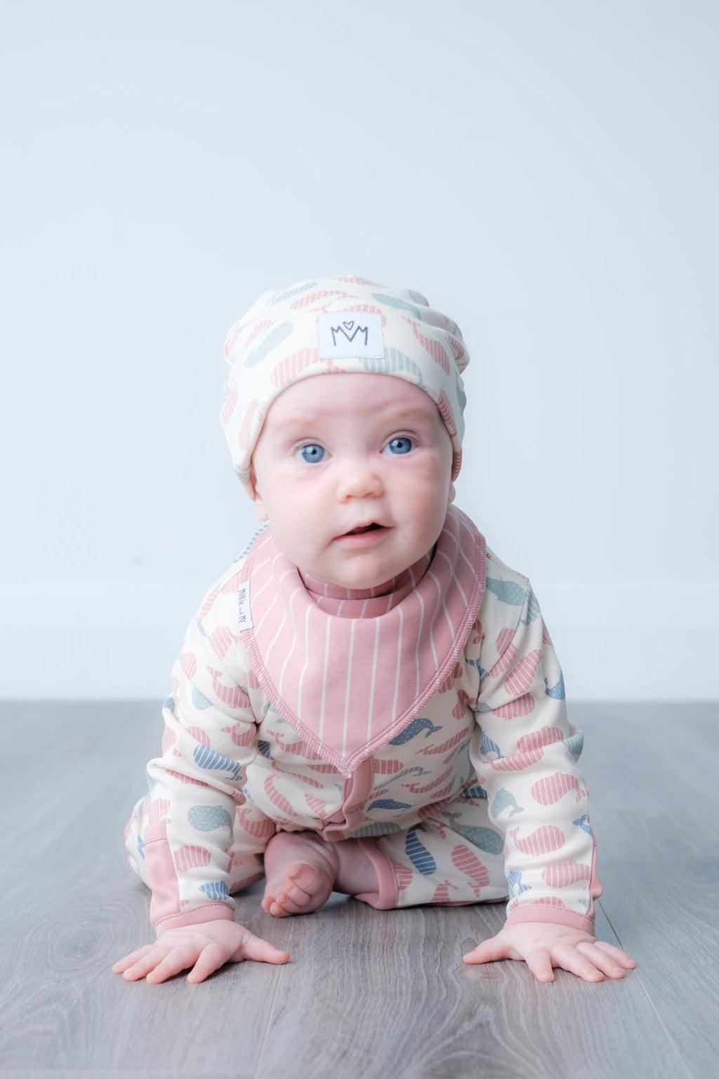 A baby wearing a pink and blue patterned long-sleeve romper, matching beanie hat, and a pink striped bib, crawling on a grey wooden floor against a light grey background.
