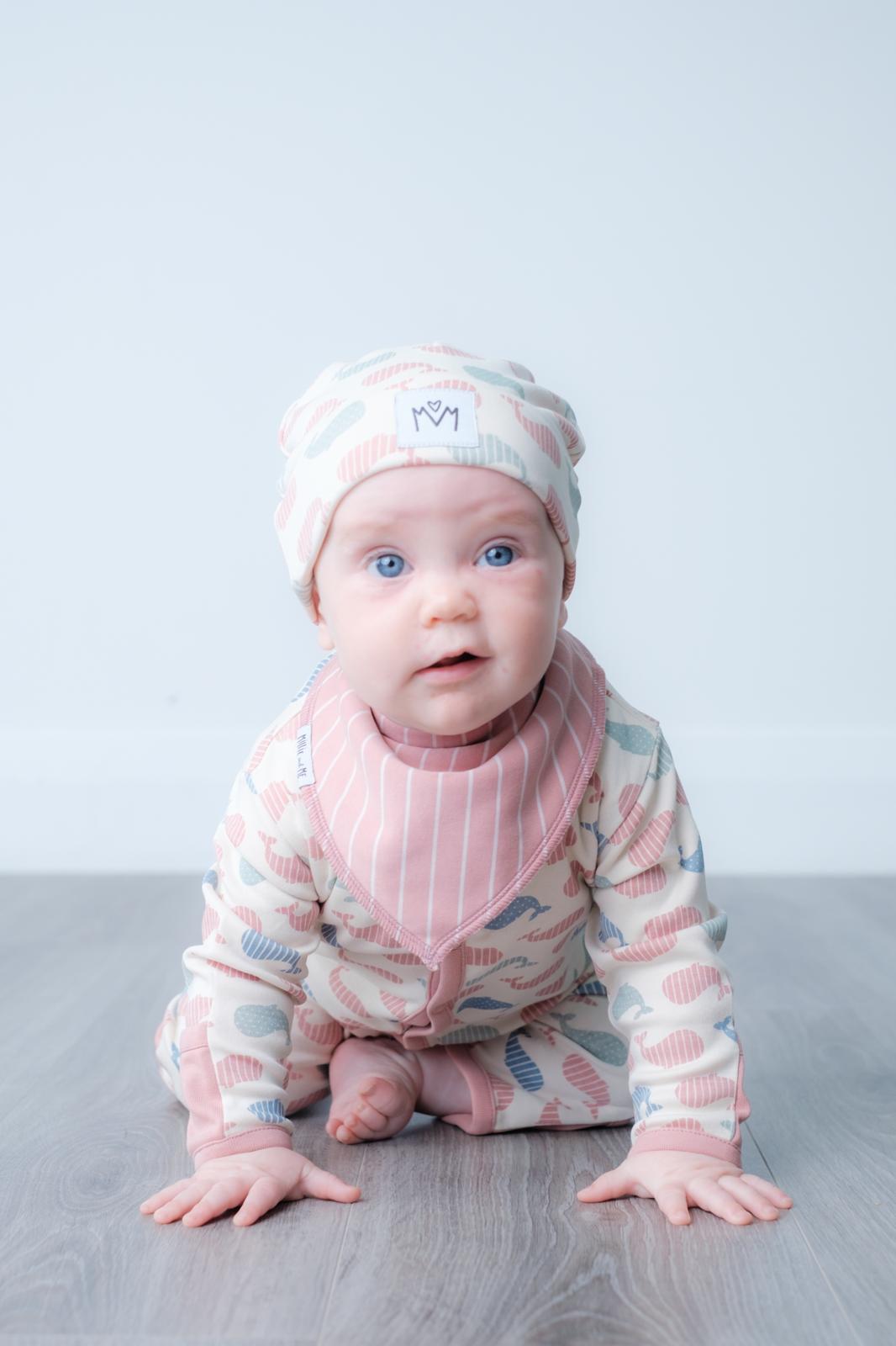 A baby wearing a pink and blue patterned long-sleeve romper, matching beanie hat, and a pink striped bib, crawling on a grey wooden floor against a light grey background.