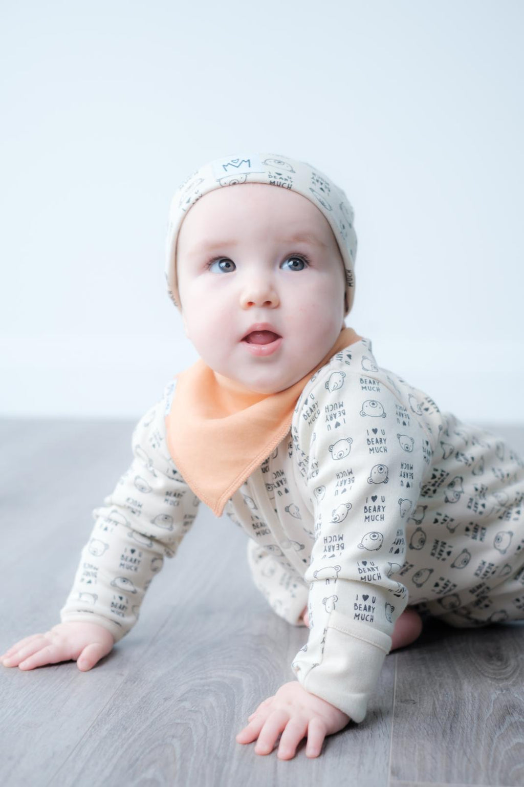 A baby crawling on a grey wood floor against a light grey background, wearing a cream-coloured long-sleeve onesie and matching bonnet with a black print, and a peach-colored bib.
