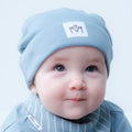 A close-up of a baby wearing a light blue knit beanie with a white square patch featuring a stylised 'MM' logo, paired with a matching blue and white striped bib.
