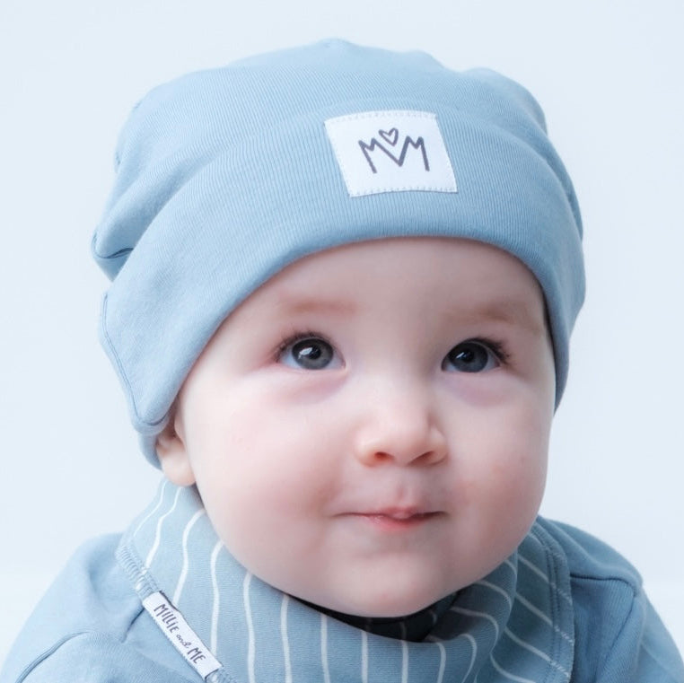 A close-up of a baby wearing a light blue knit beanie with a white square patch featuring a stylised 'MM' logo, paired with a matching blue and white striped bib.