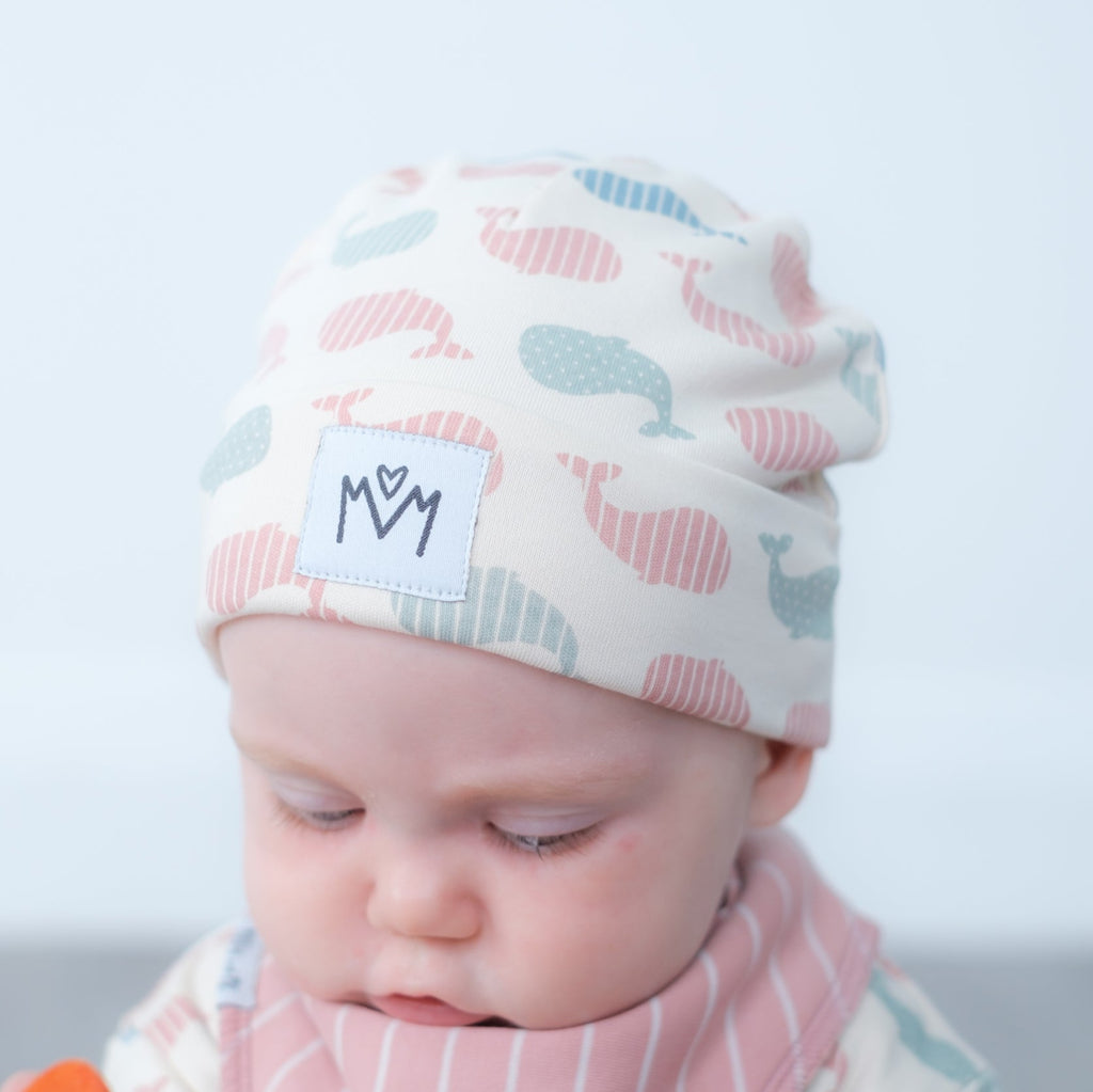 A close-up of a baby wearing a soft knit beanie hat with a whale pattern in pink, blue, and gray on an off-white background. The hat has a white patch on the front with the letters 'M' and 'M' stitched inside a heart.
