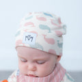A close-up of a baby wearing a soft knit beanie hat with a whale pattern in pink, blue, and gray on an off-white background. The hat has a white patch on the front with the letters 'M' and 'M' stitched inside a heart.