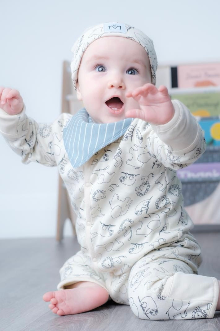 A baby wearing a cream-coloured footed onesie with a black animal print, a matching beanie, and a light blue striped bib, sitting on a wooden floor.