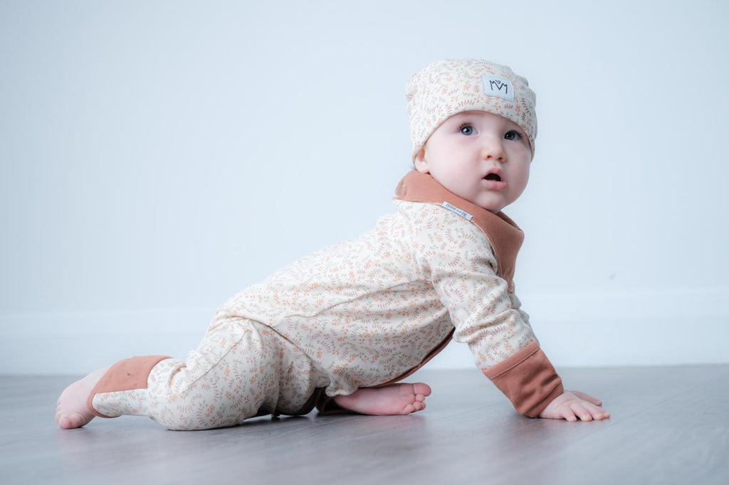 A baby crawling on a light grey floor against a pale blue-grey background, wearing a cream and tan patterned footed romper with brown cuffs and a matching knit beanie.