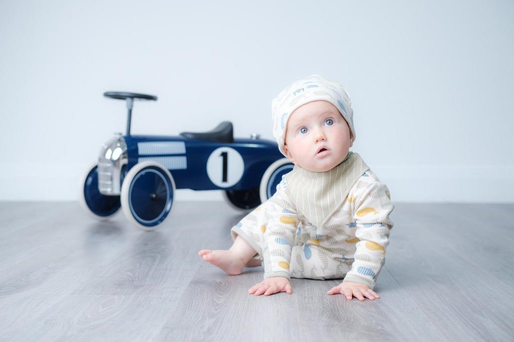 A baby wearing a cream knit outfit with yellow and blue stripes and a matching beanie sits on a gray floor, looking up at the camera. A blue toy race car with the number 1 is in the background against a light grey wall.