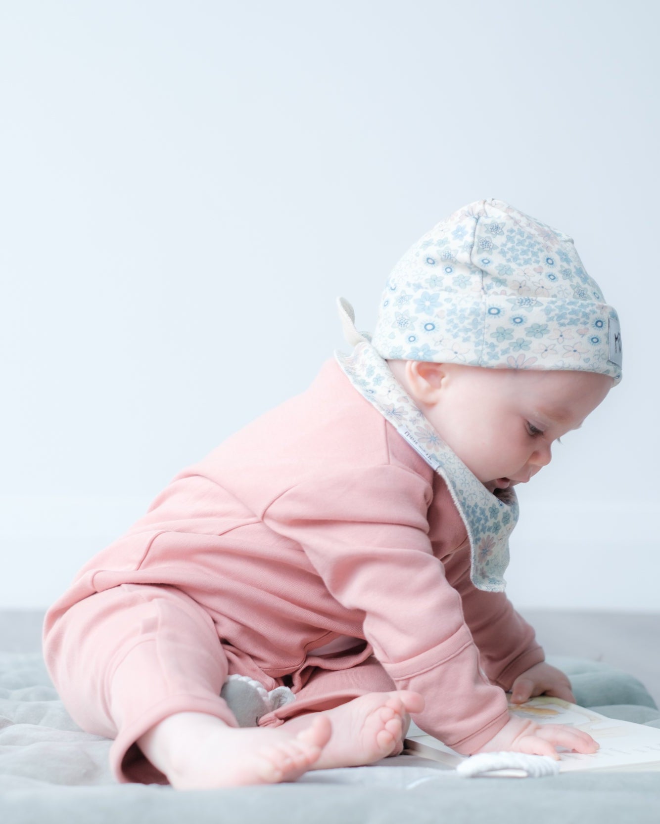 A baby wearing a pink outfit, a light blue floral bonnet, and a white bib sits on a light gray blanket on the floor, looking down at a book.