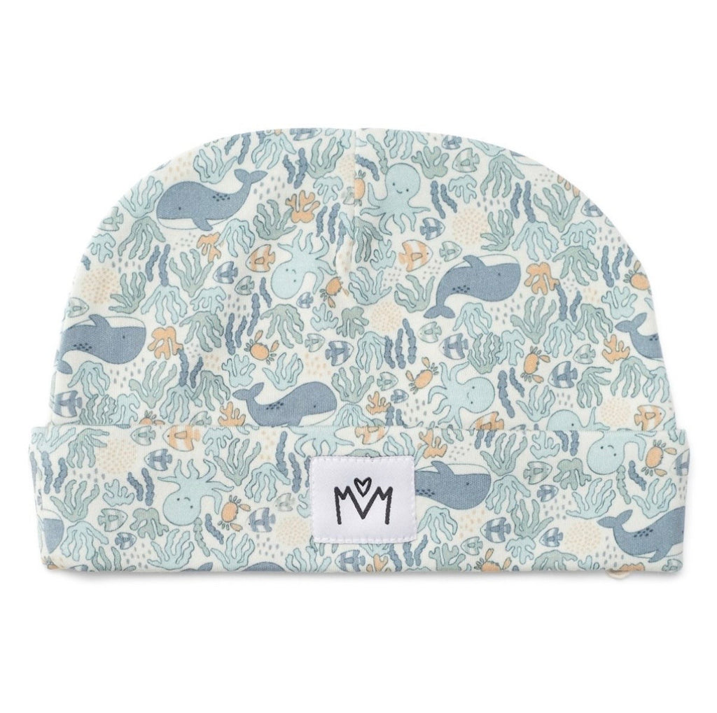 A baby beanie hat featuring a playful underwater pattern with whales, octopus, sea plants, and small fish in muted blue, gray, and beige tones. A small white fabric brand patch with a black logo is sewn onto the front cuff.
