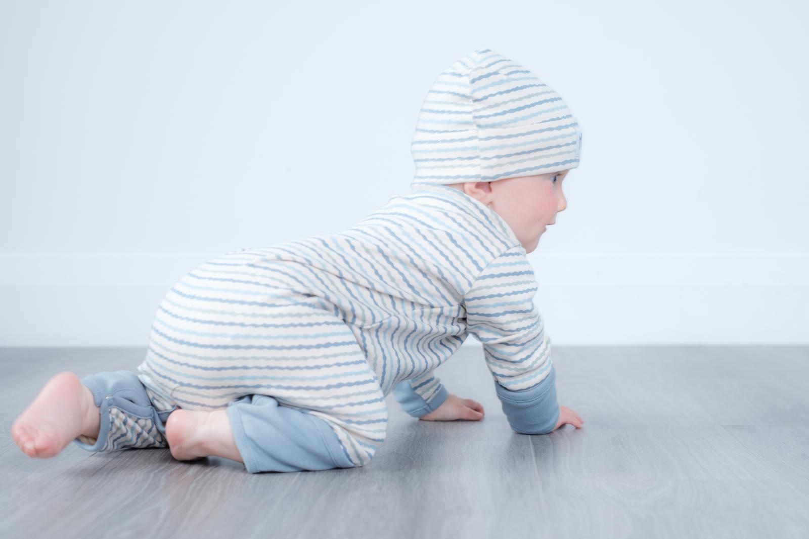 A baby crawling on a grey floor against a white wall, wearing a light blue and white striped long-sleeve romper and matching beanie hat.