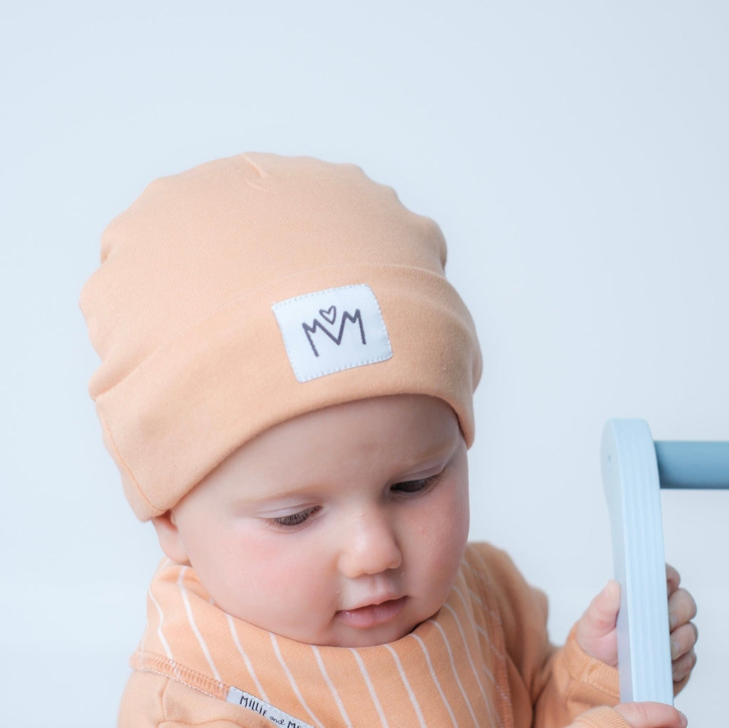 A baby wearing a peach-colored beanie with a white patch that says 'MM' and a matching striped top, holding a light blue toy against a plain light grey background.