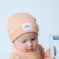 A baby wearing a peach-colored beanie with a white patch that says 'MM' and a matching striped top, holding a light blue toy against a plain light grey background.