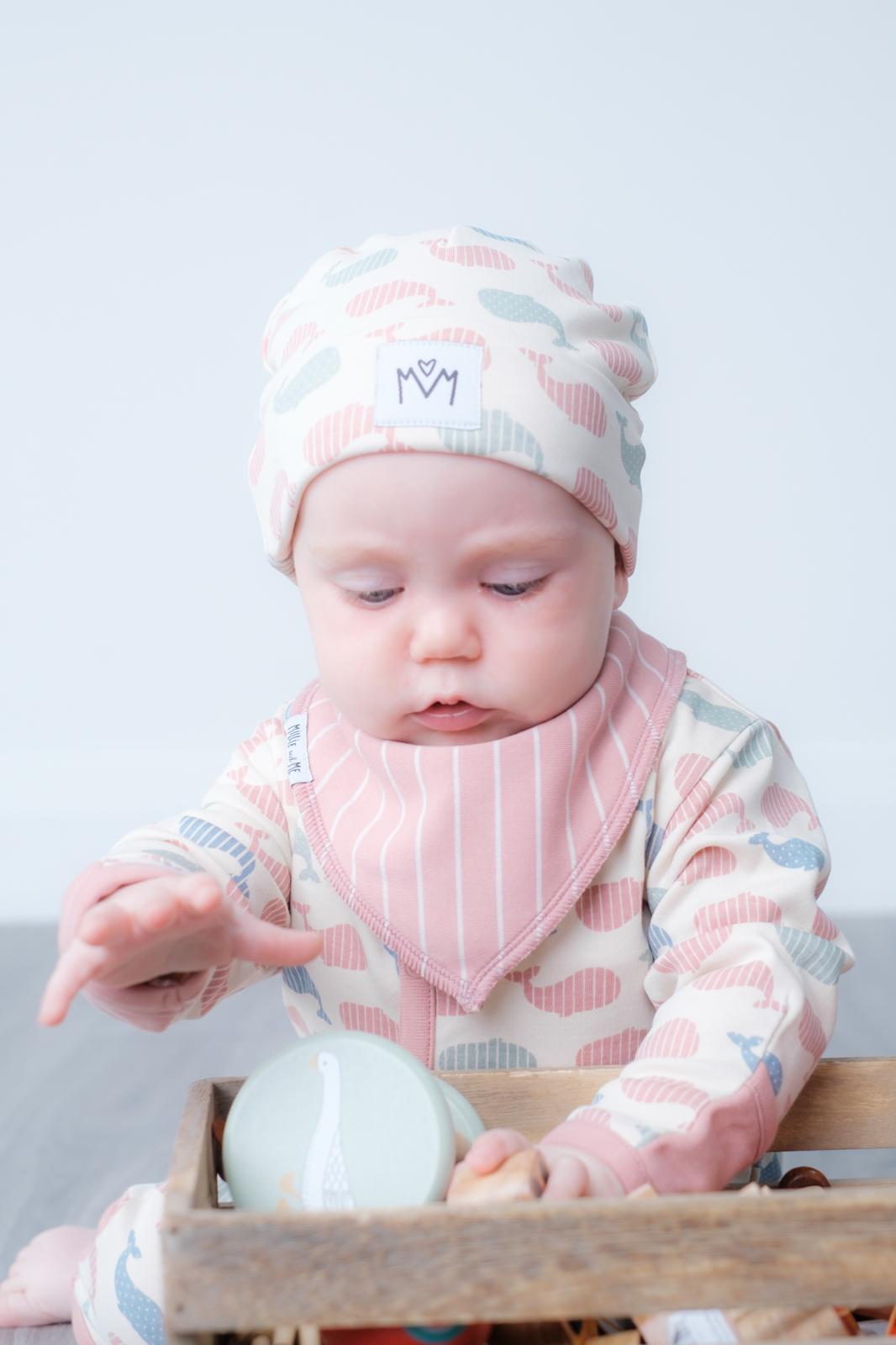 A baby wearing a soft knit hat and a pink striped bib with a matching patterned long-sleeve top, sitting in a wooden tray and reaching for a bottle.