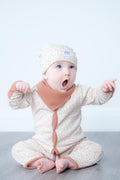 A baby wearing a beige and white patterned long-sleeve romper with brown cuffs and a matching beanie hat, sitting on a grey floor against a white wall.
