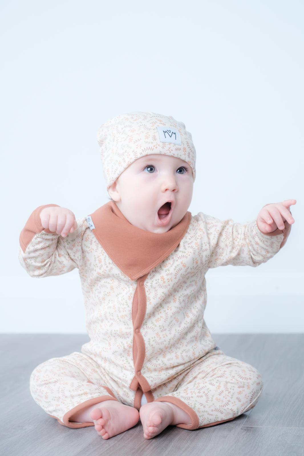 A baby wearing a beige and white patterned long-sleeve romper with brown cuffs and a matching beanie hat, sitting on a grey floor against a white wall.