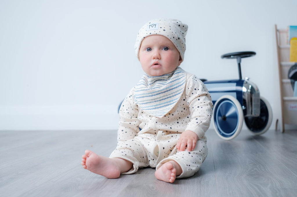 A baby sitting on a grey wooden floor in a nursery, wearing a cream-colored long-sleeve romper with tiny dark polka dots, a matching beanie, and a striped bib.