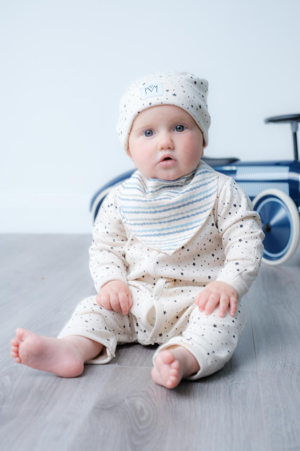 A baby sitting on a grey wooden floor wearing a cream-coloured long-sleeve romper with tiny dark stars, a matching speckled bonnet, and a blue-and-white striped bib. A blue toy car is blurred in the background.