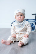 A baby sitting on a grey wooden floor wearing a cream-coloured long-sleeve romper with tiny dark stars, a matching speckled bonnet, and a blue-and-white striped bib. A blue toy car is blurred in the background.