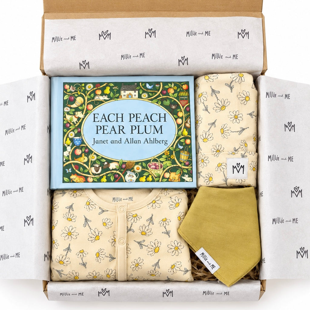 A curated baby gift set in a cardboard box from Millie and Me, featuring a children's book titled 'Each Peach Pear Plum' by Janet and Allan Ahlberg, a cream floral baby babygrow, a matching floral baby hat, and a mustard yellow bandana bib. The items are neatly arranged on wood shavings, inside tissue-wrapped packaging.