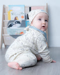 A baby wearing a cream-colored long-sleeve romper and matching beanie hat with a gray animal print, crawling on a gray wood-look floor in front of a light-colored bookshelf.