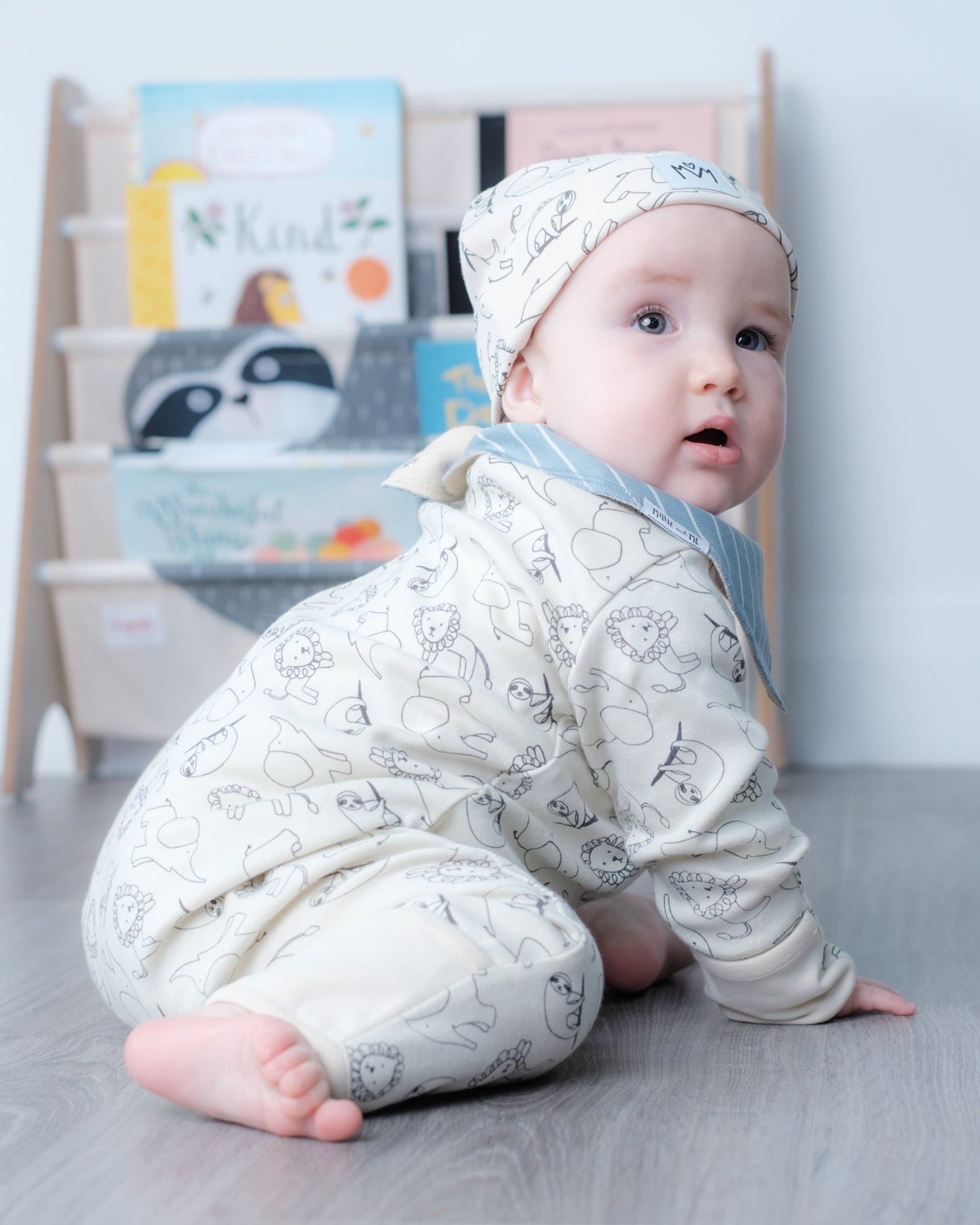A baby wearing a cream-colored long-sleeve romper and matching beanie hat with a gray animal print, crawling on a gray wood-look floor in front of a light-colored bookshelf.