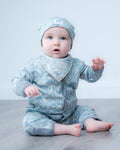 A baby sitting on a gray wood floor against a white wall, wearing a light blue patterned one-piece outfit with a matching hat and bib.