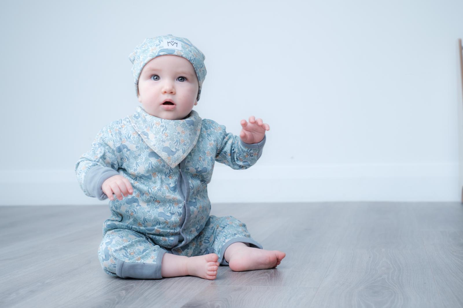A baby sitting on a gray wood floor against a white wall, wearing a light blue patterned one-piece outfit with a matching hat and bib.