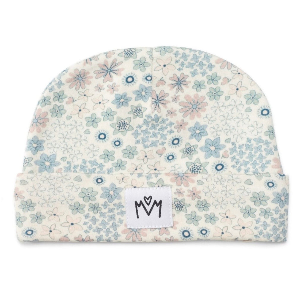 A folded baby beanie hat featuring a delicate floral pattern in shades of blue, pink, and green on a warm cream background. A small white square patch with a black hand-drawn logo is sewn onto the front cuff.