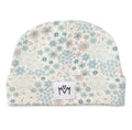 A folded baby beanie hat featuring a delicate floral pattern in shades of blue, pink, and green on a warm cream background. A small white square patch with a black hand-drawn logo is sewn onto the front cuff.