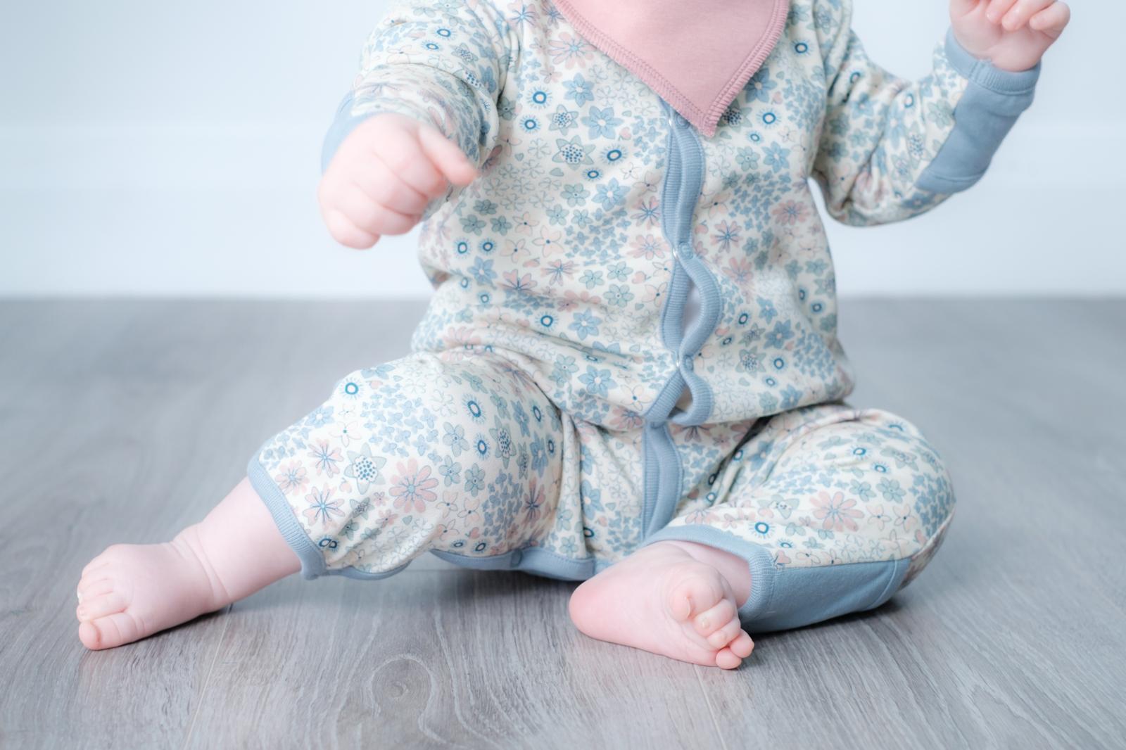 A baby sitting on a gray wood floor wearing a light blue and pink floral long-sleeve footless sleeper with a pink bib.